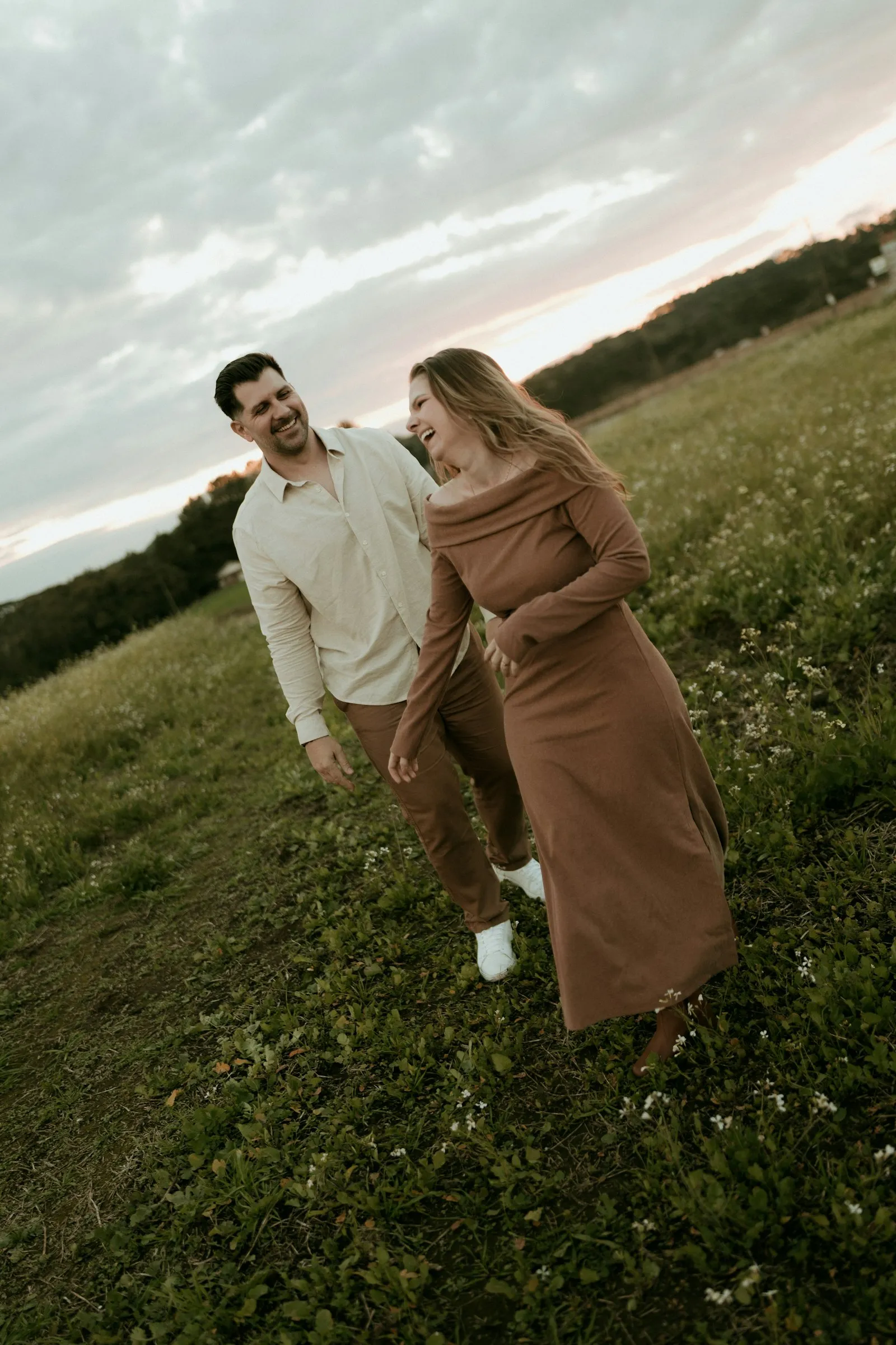 A couple laughing together as they walk through a meadow at sunset